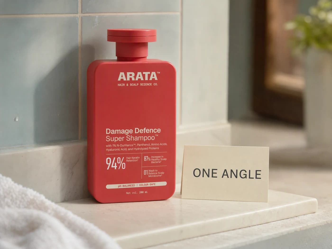 A red Arata shampoo bottle on a light stone bathroom shelf in daylight beside a small cream card reading one angle, with a folded white towel behind it.