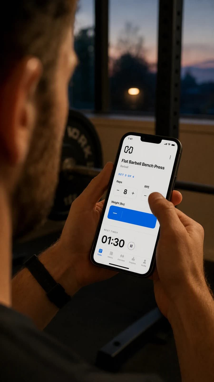 An over-the-shoulder view of a lifter's thumb mid-tap on a workout-logging screen at dusk, the weight field just out of frame.