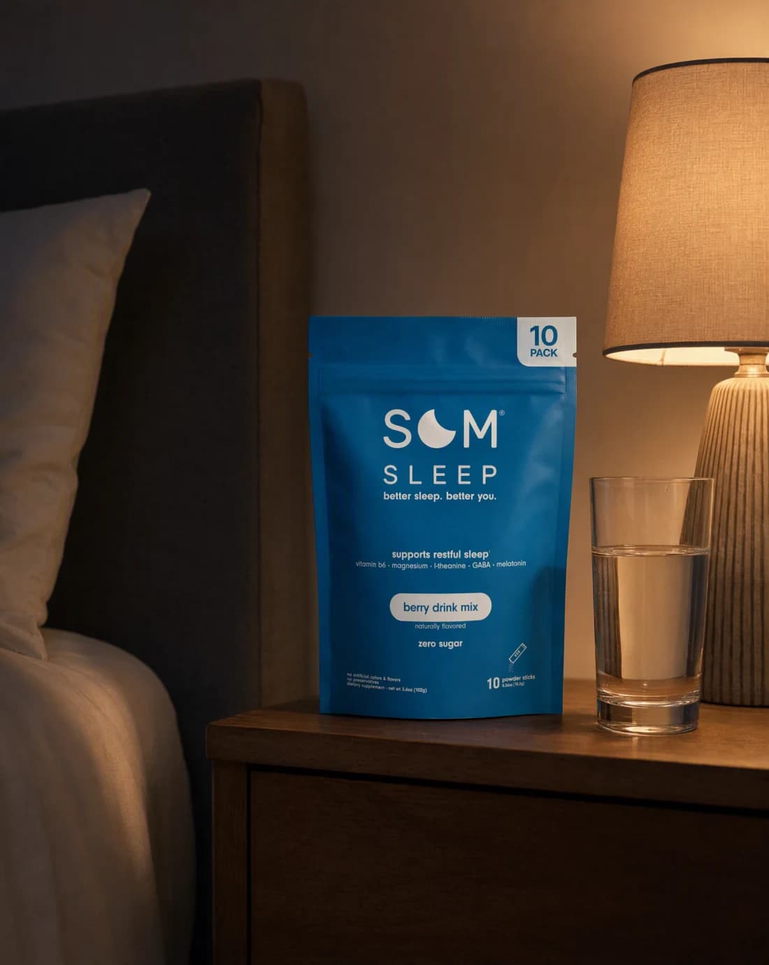 A Som Sleep drink mix pack on a bedside table beside a glass of water, lit by a small warm lamp at night.
