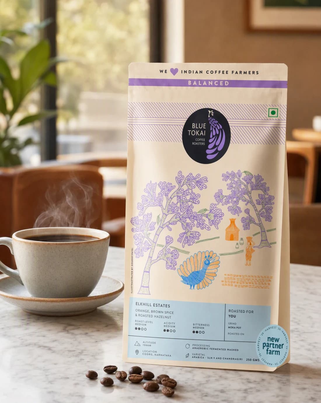 A Blue Tokai coffee bag standing on a marble cafe counter beside a steaming ceramic cup and a scatter of whole coffee beans.