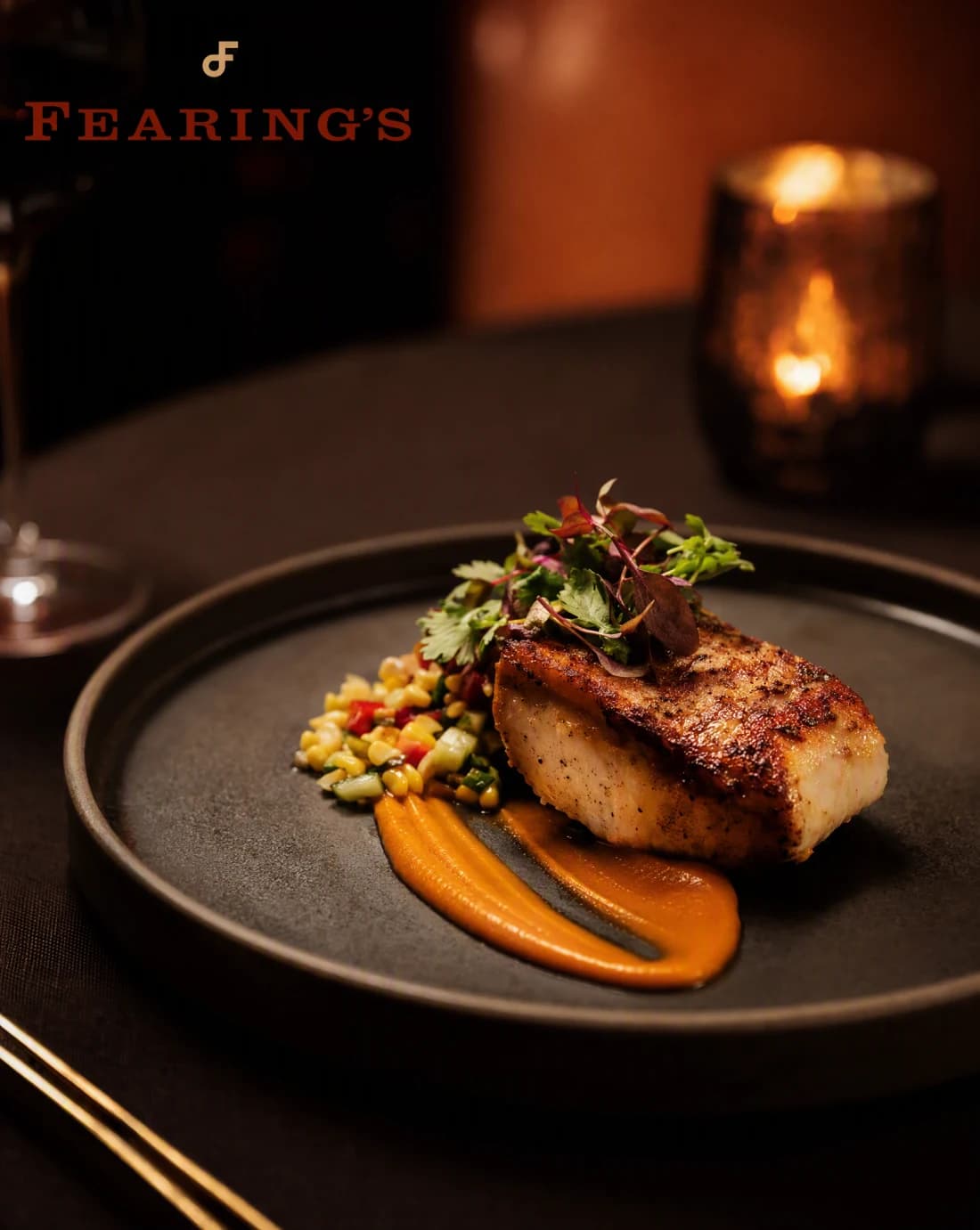 A seared, plated dish on a dark ceramic plate at a restaurant table, warm low light and a candle glowing softly out of focus behind it.