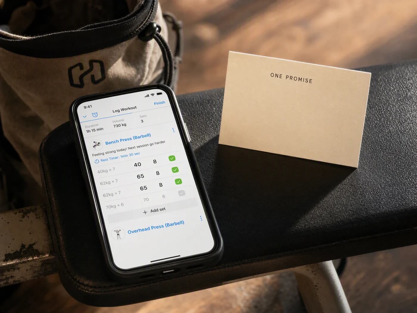 A phone on a gym bench showing the Hevy workout logging screen, beside an upright cream card reading one promise, with a chalk bag blurred behind it.