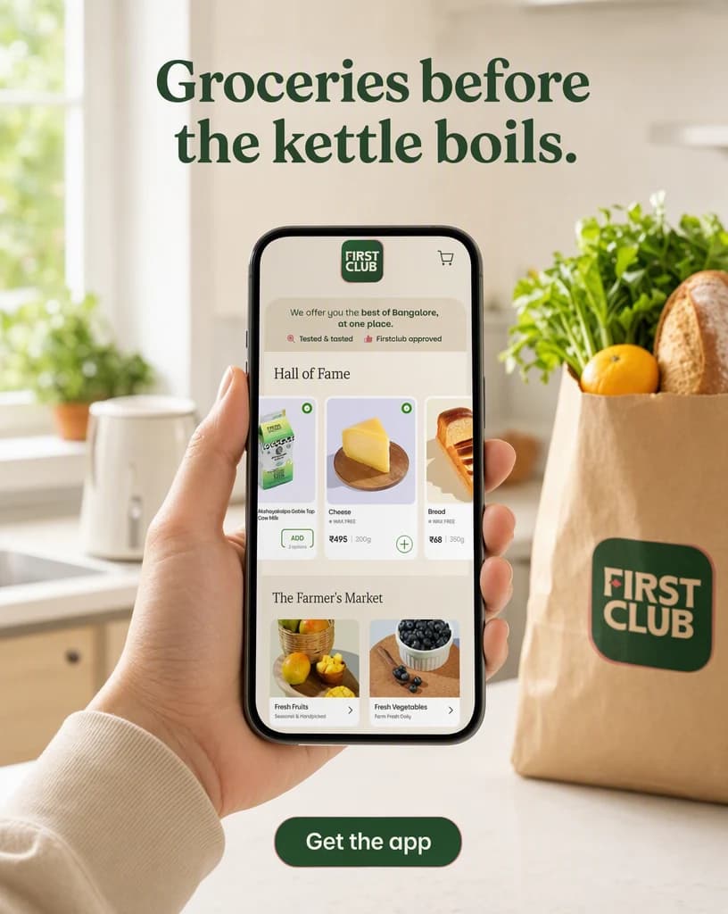 A hand holding a phone showing the FirstClub app screen in a bright kitchen with a paper bag of groceries on the counter behind, under a headline about groceries arriving before the kettle boils.