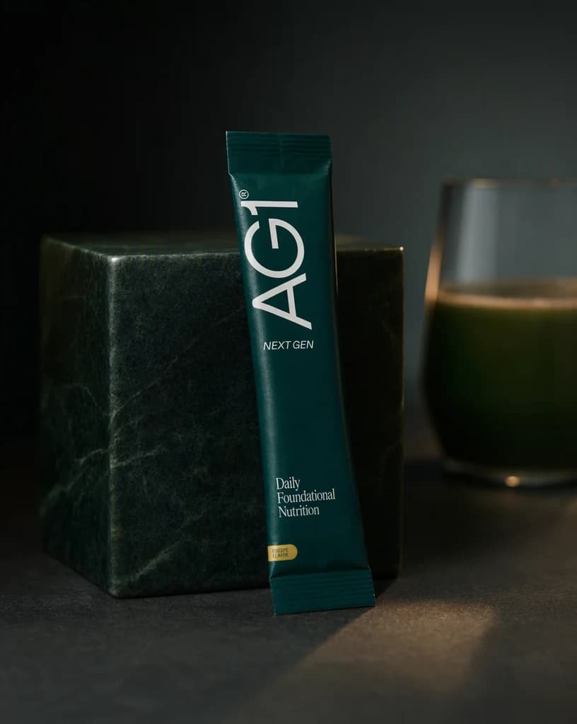 An AG1 travel pack sachet propped against a smooth dark green stone block on a matte charcoal surface, a glass of green drink out of focus behind it.